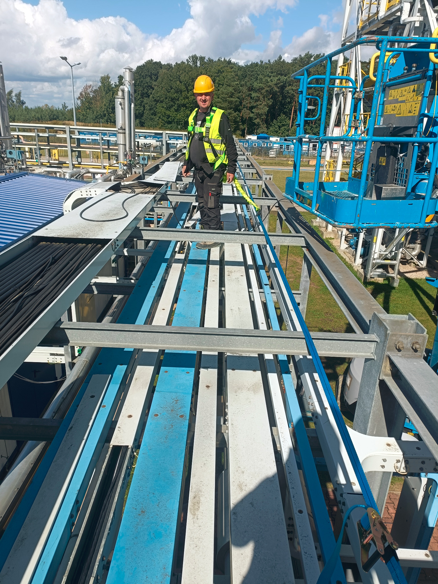 Pracownik w kasku i kamizelce na konstrukcji stalowej z kablami, prawdopodobnie podczas prac konserwacyjnych lub montażowych na terenie przemysłowym.