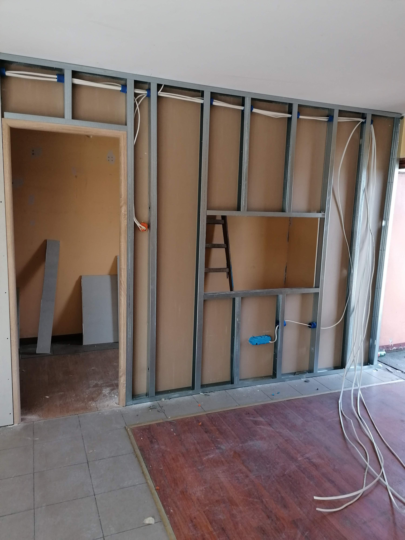 Montaż ścianki działowej z instalacją elektryczną wewnątrz. Widoczne kable, metalowa konstrukcja i płyty kartonowo-gipsowe. W tle drabina i fragment pomieszczenia.