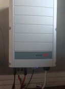 Biała obudowa falownika fotowoltaicznego SMA z widocznymi kablami u dołu, zamontowana na jasnej ścianie. Minimalistyczny design, lekki kąt ujęcia.