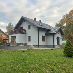 Profmontaż ihor lenchenko - Nowoczesny dom jednorodzinny z jasną elewacją, ciemną dachówką i drewnianymi elementami balkonu, otoczony zielonym trawnikiem i jesiennymi drzewami.