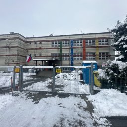 Profmontaż ihor lenchenko - Budynek w rusztowaniach, częściowo ocieplony, z flagą Polski i UE. Zima, śnieg. Kolorowe pasy na elewacji. Tabliczki ostrzegawcze na ogrodzeniu.