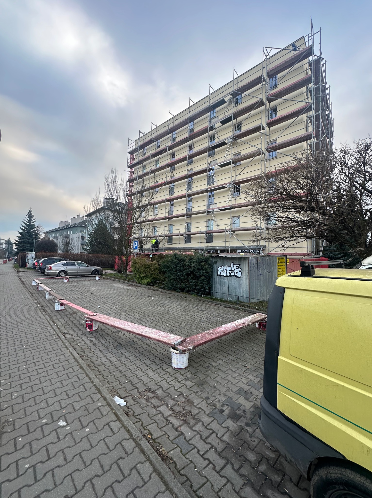 Wielopiętrowy budynek w trakcie ocieplania, otoczony rusztowaniami. Widoczne otwarte puszki z farbą i fragment żółtego samochodu dostawczego. Pochmurne niebo.