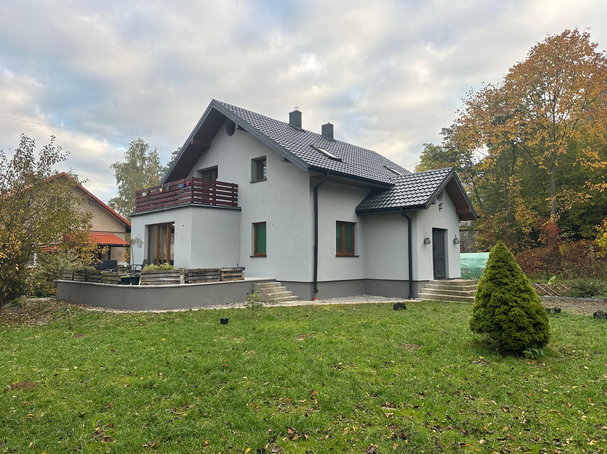 Nowoczesny dom jednorodzinny z jasną elewacją, ciemną dachówką i drewnianymi elementami balkonu, otoczony zielonym trawnikiem i jesiennymi drzewami.