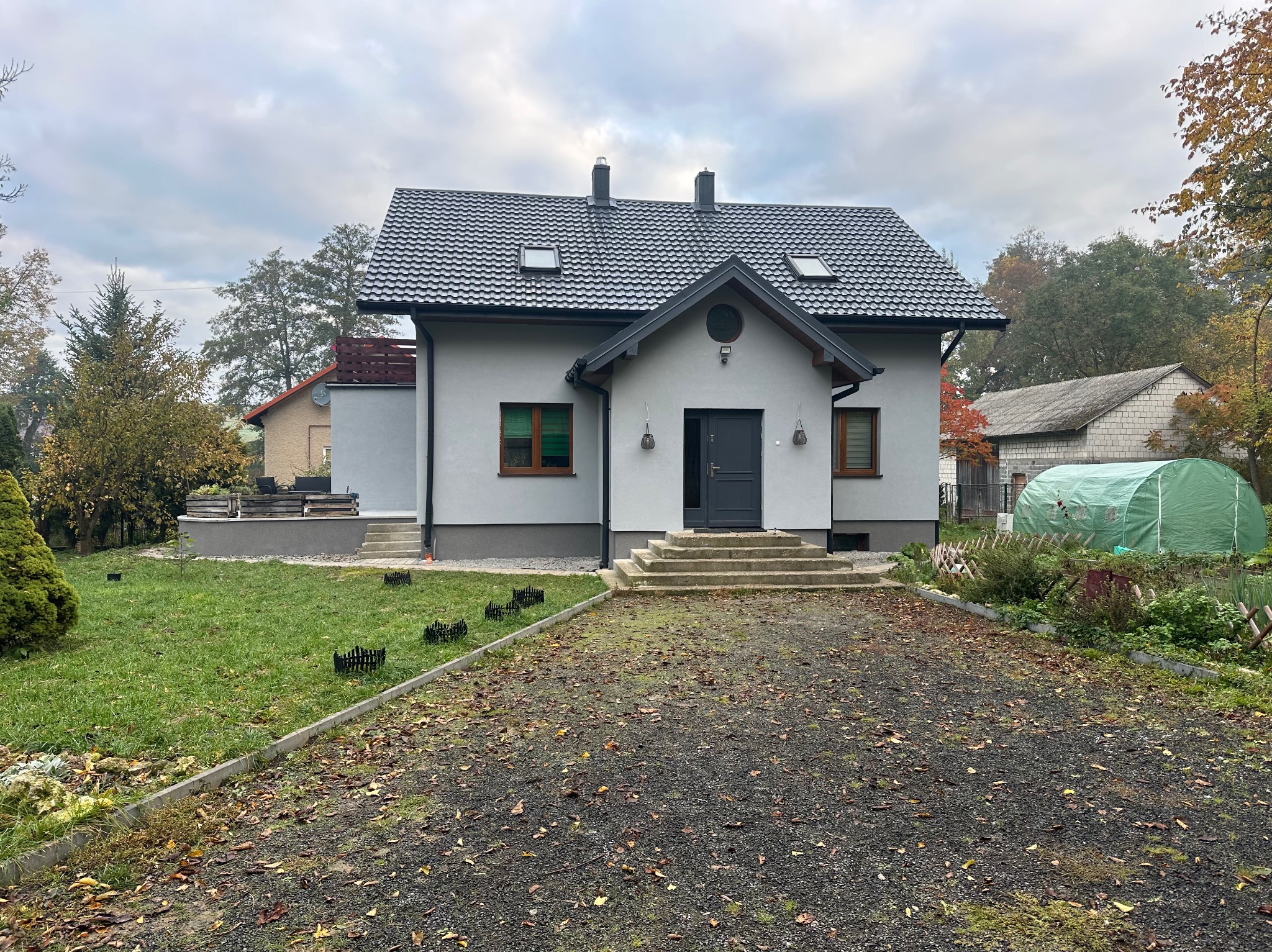 Jasnoszary dom jednorodzinny z ciemnoszarą stolarką i dachem z dachówki, widok z przodu, jesienne otoczenie. Widoczne detale wykończenia elewacji.