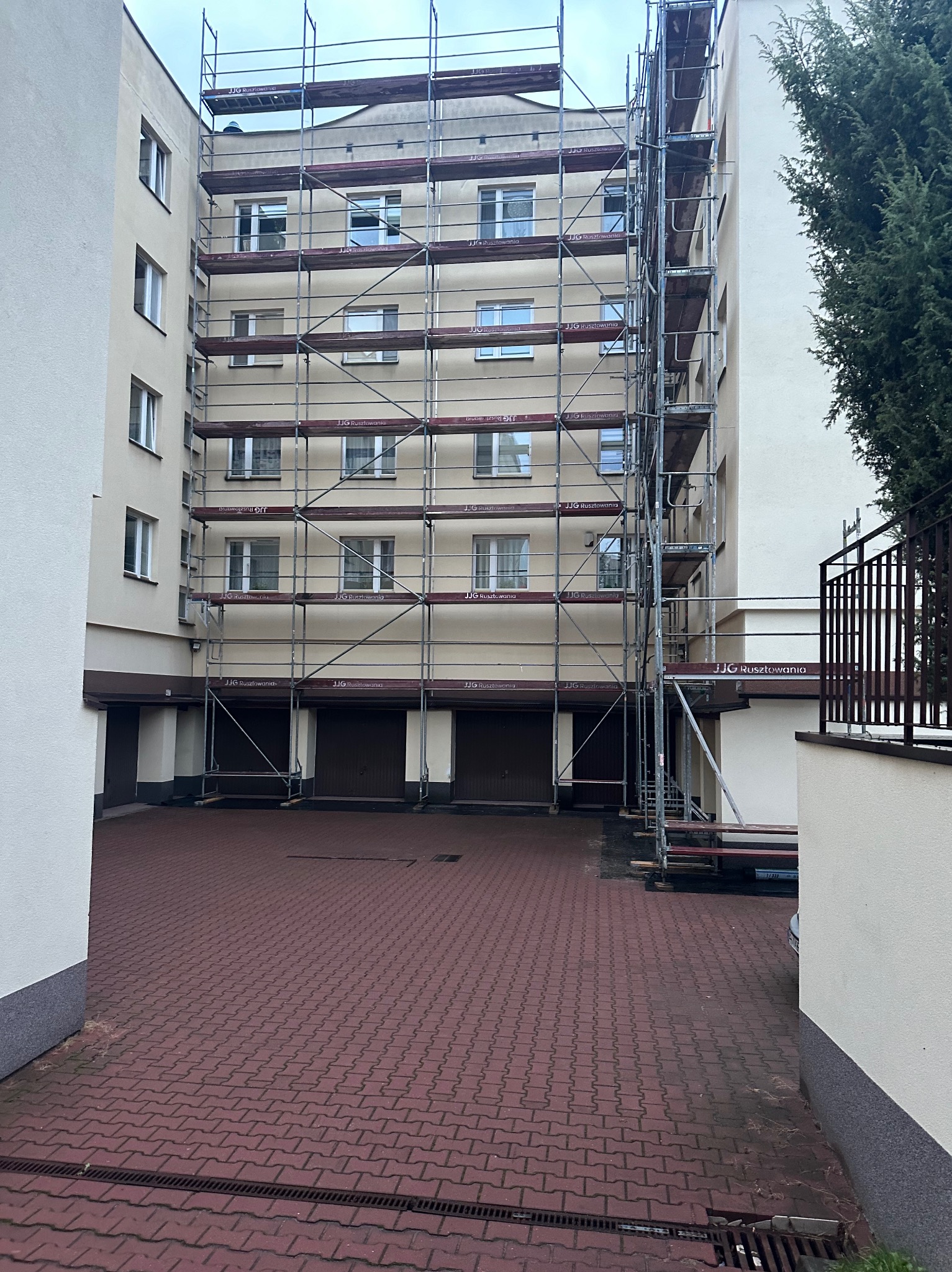Budynek wielorodzinny w trakcie prac elewacyjnych, osłonięty rusztowaniem z widocznymi oznaczeniami producenta. Czerwona kostka brukowa na dziedzińcu.