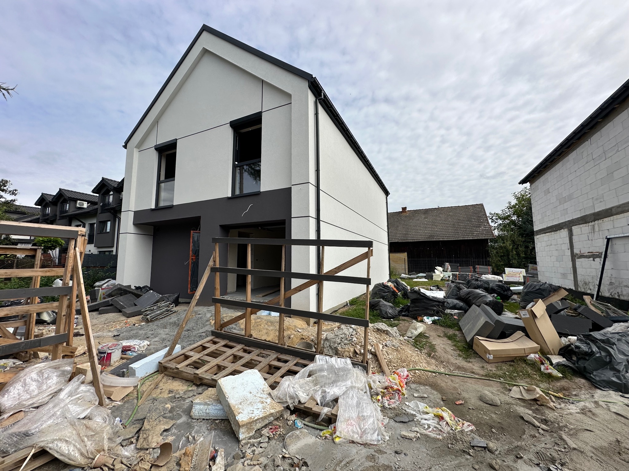 Nowoczesny dom z białą elewacją w trakcie wykończenia. Widoczne rusztowania i materiały budowlane. Uwagę zwraca minimalistyczny design i dwukolorowa fasada.