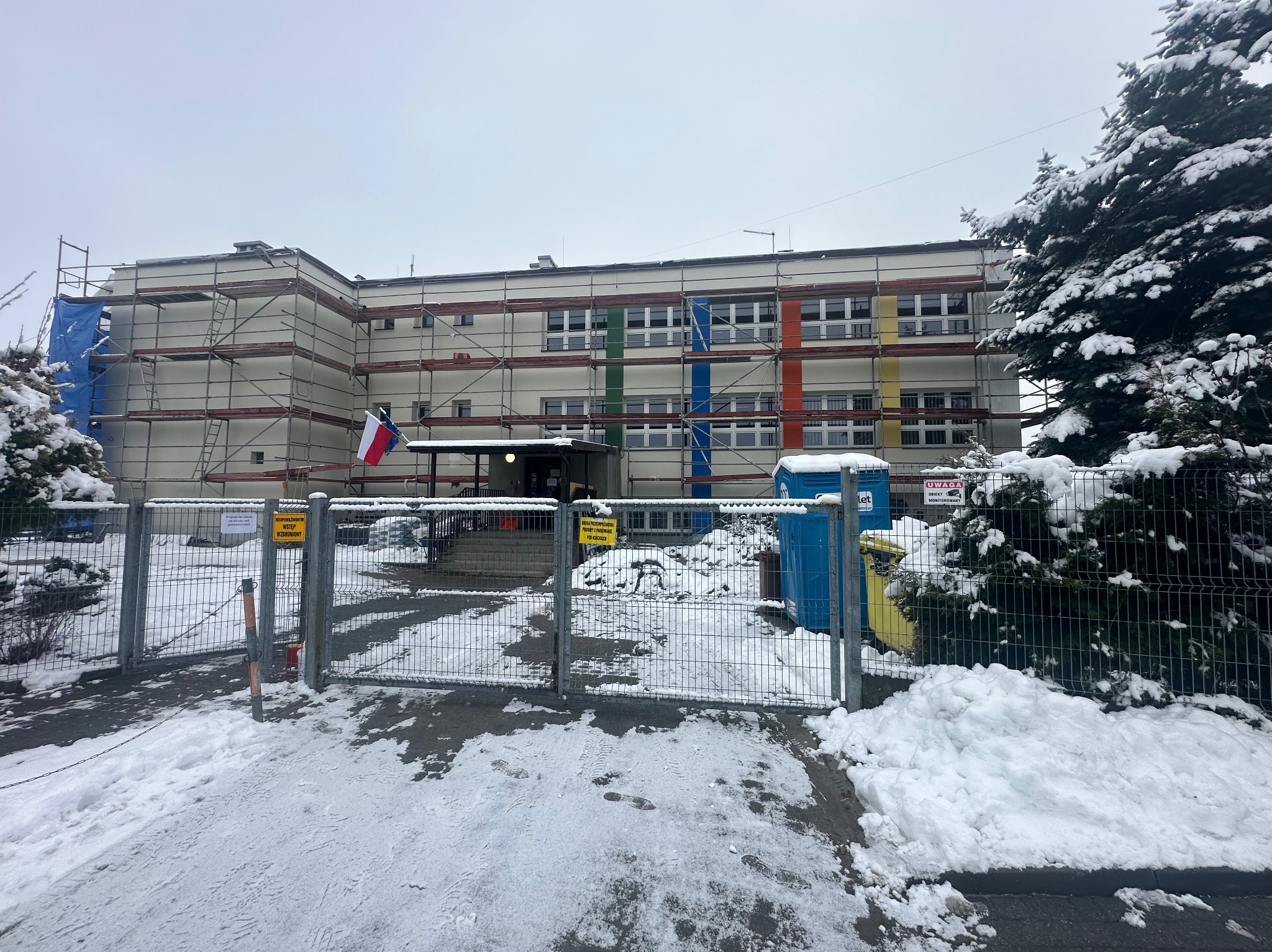 Budynek w rusztowaniach, częściowo ocieplony, z flagą Polski i UE. Zima, śnieg. Kolorowe pasy na elewacji. Tabliczki ostrzegawcze na ogrodzeniu.
