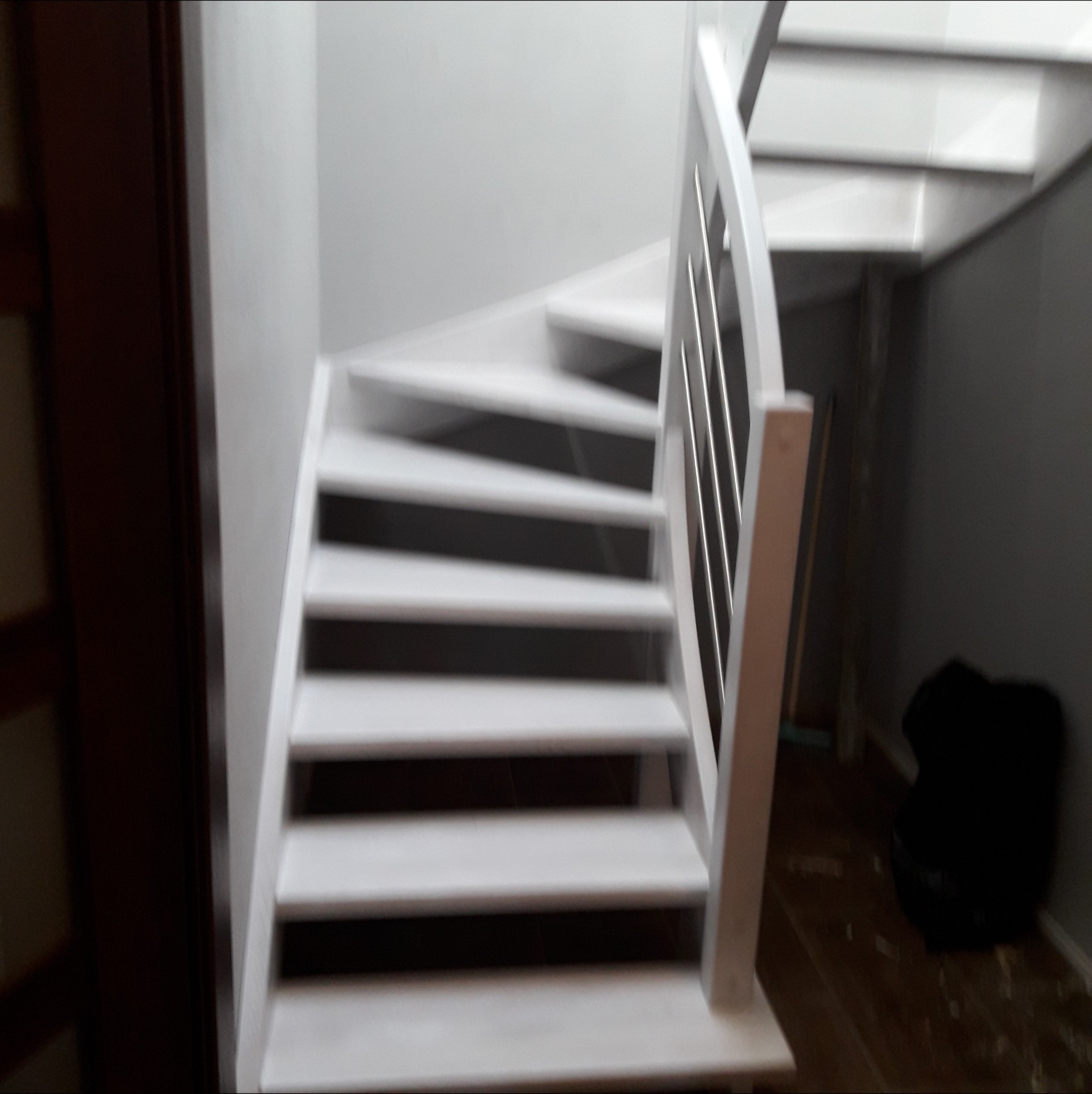 Białe drewniane schody zabiegowe z metalowymi prętami balustrady, widoczne z dołu, z ciemnym tłem i fragmentem drzwi po lewej stronie.