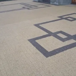 Ułożona jasna kostka brukowa z wzorem geometrycznym z ciemniejszej kostki. Widok z góry, pokazujący fragment chodnika z dekoracyjnym wzorem.