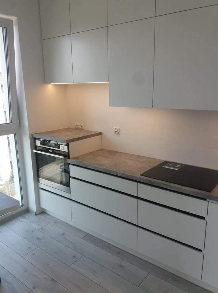 Nowoczesna kuchnia z białymi, minimalistycznymi frontami szafek i szarym blatem z subtelnym wzorem, płyta indukcyjna wbudowana w blat, oświetlenie podszafkowe LED.