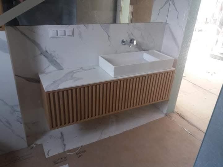 Łazienkowa szafka pod umywalkę z frontem z pionowych listewek w kolorze jasnego drewna, biała umywalka nablatowa, tło z płytek imitujących marmur, widoczne przyłącza baterii.