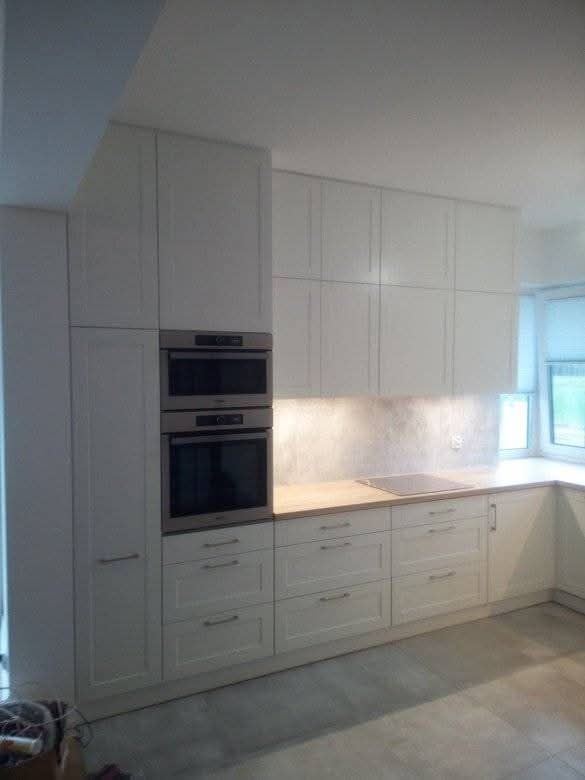 Jasna kuchnia z białymi frontami szafek i drewnianym blatem, zabudowana pod sufit z wbudowanym piekarnikiem i płytą grzewczą, oświetlona od dołu.