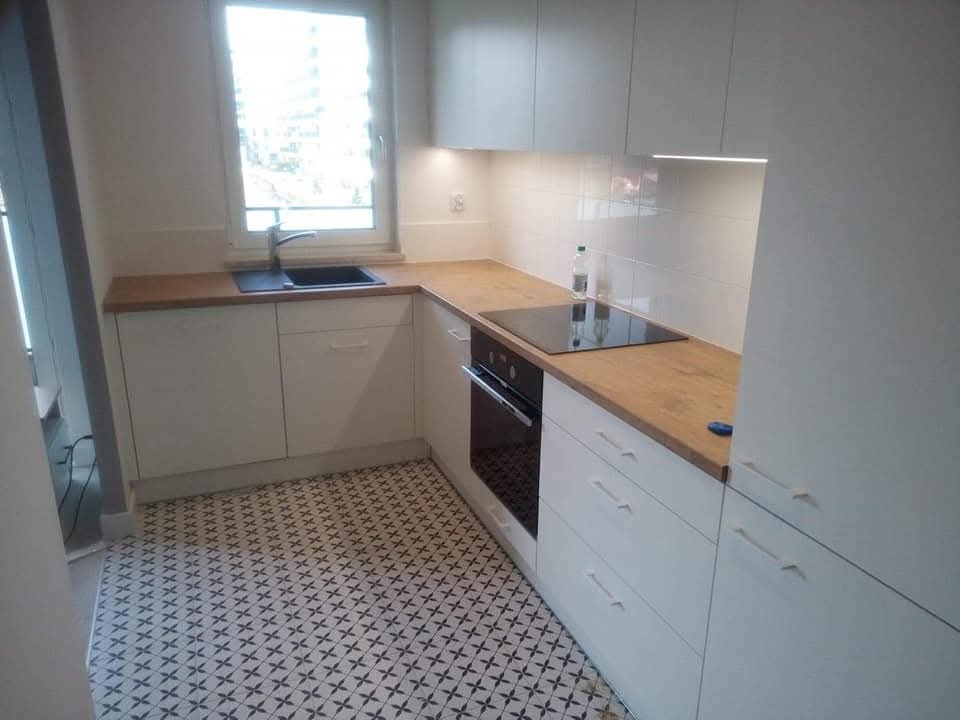 Jasna kuchnia z białymi szafkami i drewnianym blatem, czarny zlew pod oknem i płyta indukcyjna, podświetlenie LED pod szafkami wiszącymi, wzorzysta podłoga w czarno-białe gwiazdki.