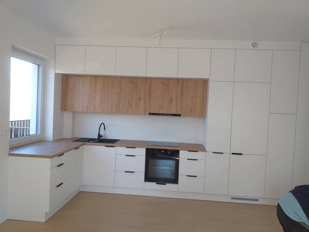 Nowoczesna, jasna kuchnia z białymi szafkami i drewnianymi akcentami, czarnym zlewem i blatem w odcieniu drewna, minimalistyczne uchwyty.