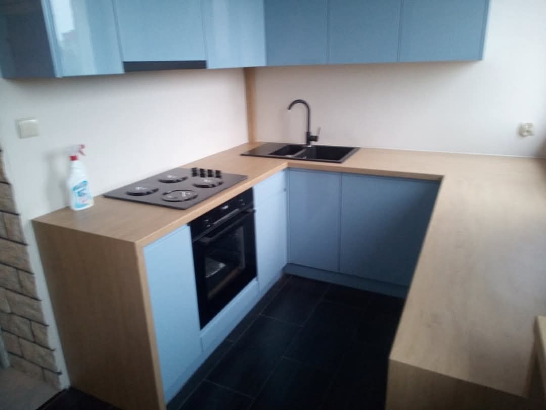 Jasna kuchnia w kształcie litery U z błękitnymi szafkami, czarnym zlewem i płytą grzewczą, wykończona blatem w kolorze jasnego drewna, z butelką środka czyszczącego na blacie.