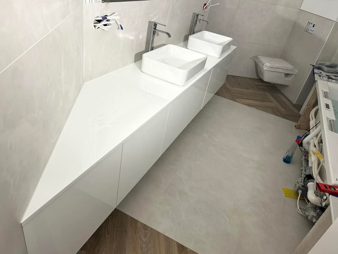 Biała, nowoczesna umywalka nablatowa z szafką podumywalkową w trakcie montażu, widoczne przewody i narzędzia, płytki w odcieniach szarości, fragment podłogi z parkietem ułożonym w jodełkę.