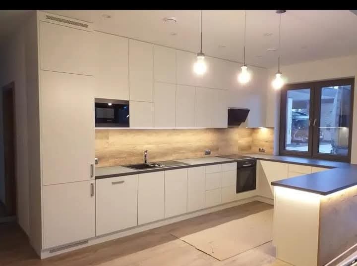 Nowoczesna, jasna kuchnia z białymi frontami, czarnym blatem i drewnopodobną ścianą nad blatem, oświetlona wiszącymi lampami i listwą LED pod szafkami górnymi; na podłodze karton zabezpieczający.