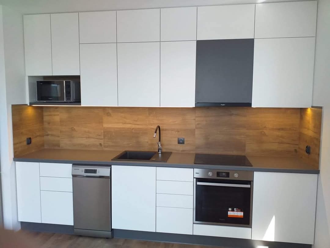 Nowoczesna kuchnia z białymi frontami szafek, drewnopodobnym panelem ściennym, szarym blatem i wbudowanym sprzętem AGD: piekarnikiem, płytą indukcyjną, zmywarką i mikrofalówką.