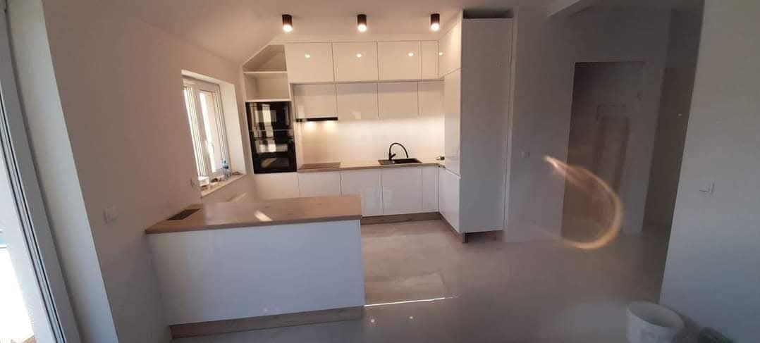 Biała, lakierowana kuchnia z drewnianym blatem, nowoczesne oświetlenie punktowe, widok z perspektywy wejścia do pomieszczenia, w tle okno.