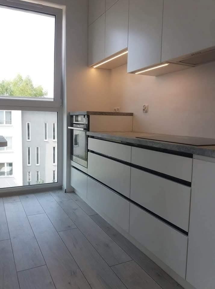 Nowoczesna kuchnia z białymi frontami szafek i szuflad z czarnymi uchwytami, szarym blatem i oknem z widokiem na budynek mieszkalny