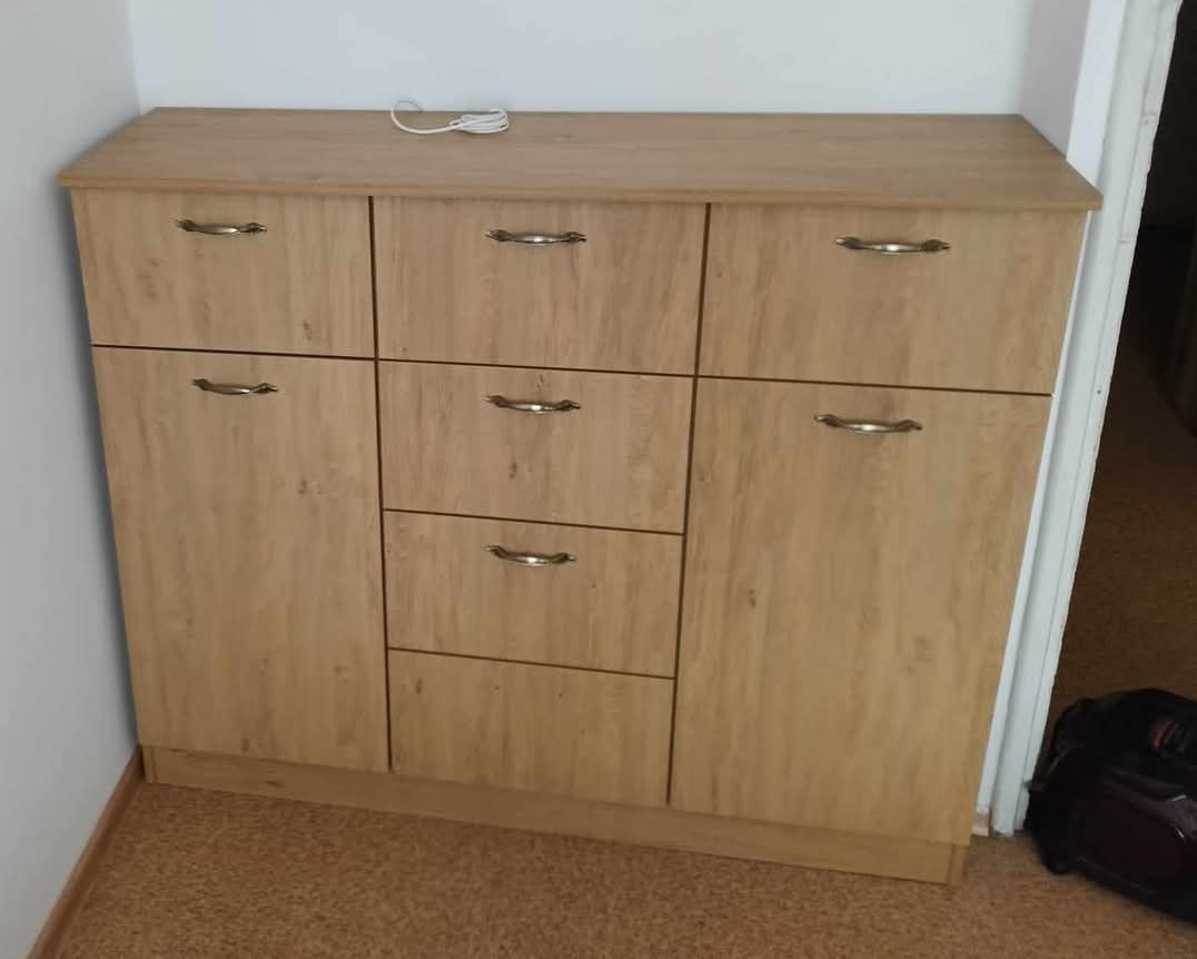 Jasna, drewnopodobna szafka z sześcioma frontami, w tym trzema szufladami, z metalowymi uchwytami, stojąca przy ścianie z widocznym kablem na blacie.