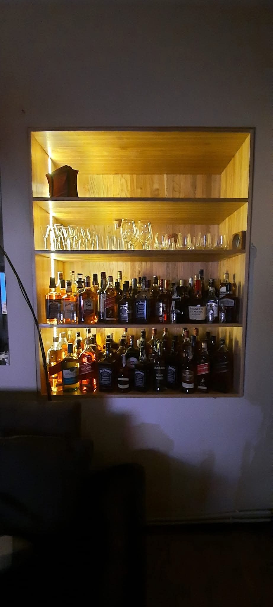 Wnękowa, podświetlana półka z jasnego drewna, wypełniona kolekcją butelek alkoholu i szkła barowego.