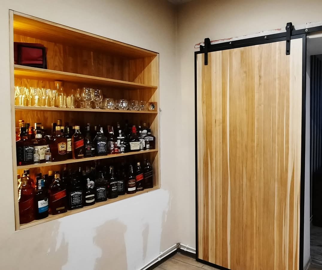 Wnęka barowa z drewnianymi półkami wypełniona butelkami alkoholu i szklankami, obok przesuwne drzwi z jasnego drewna na czarnych szynach.