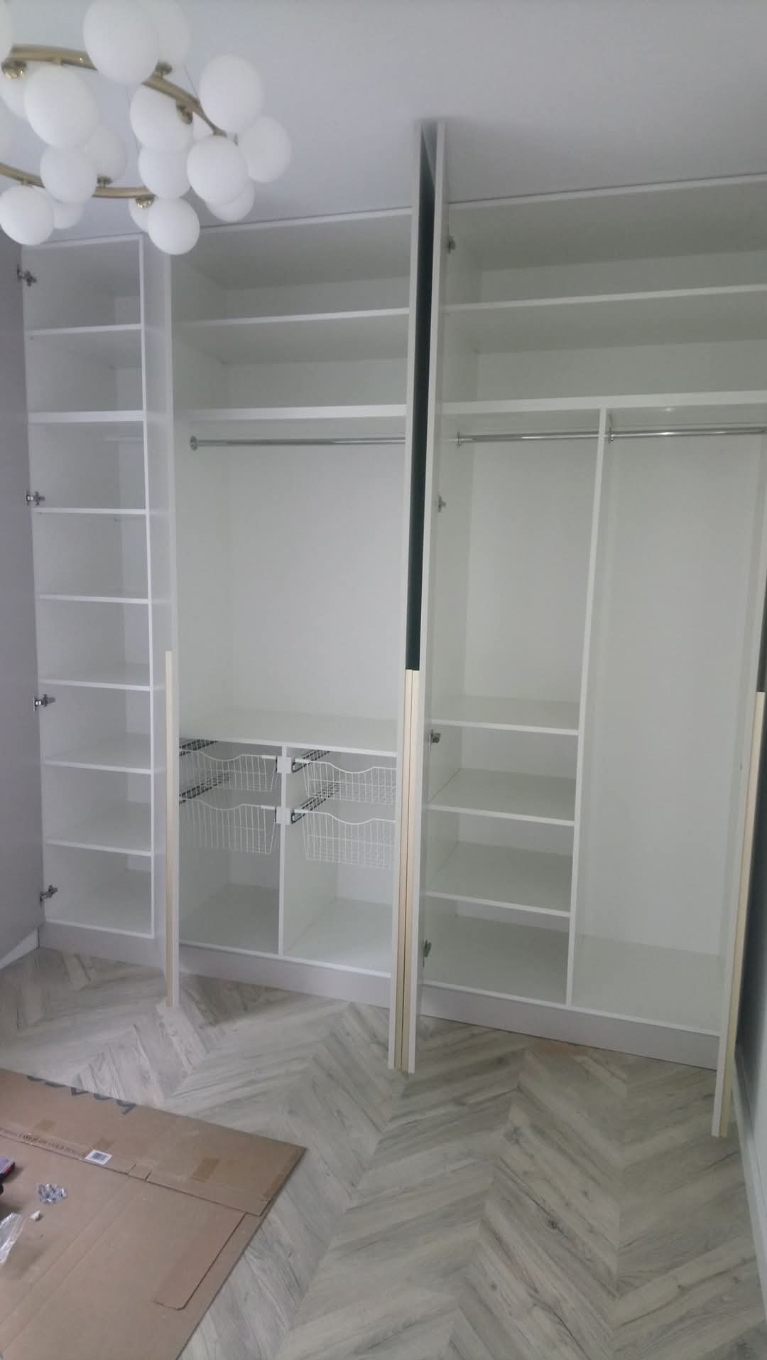 Biała, trzydrzwiowa szafa z otwartymi drzwiami, widoczne półki, drążki na ubrania i wysuwane kosze druciane, parkiet ułożony w jodełkę na podłodze, lampa sufitowa w kształcie kuli.