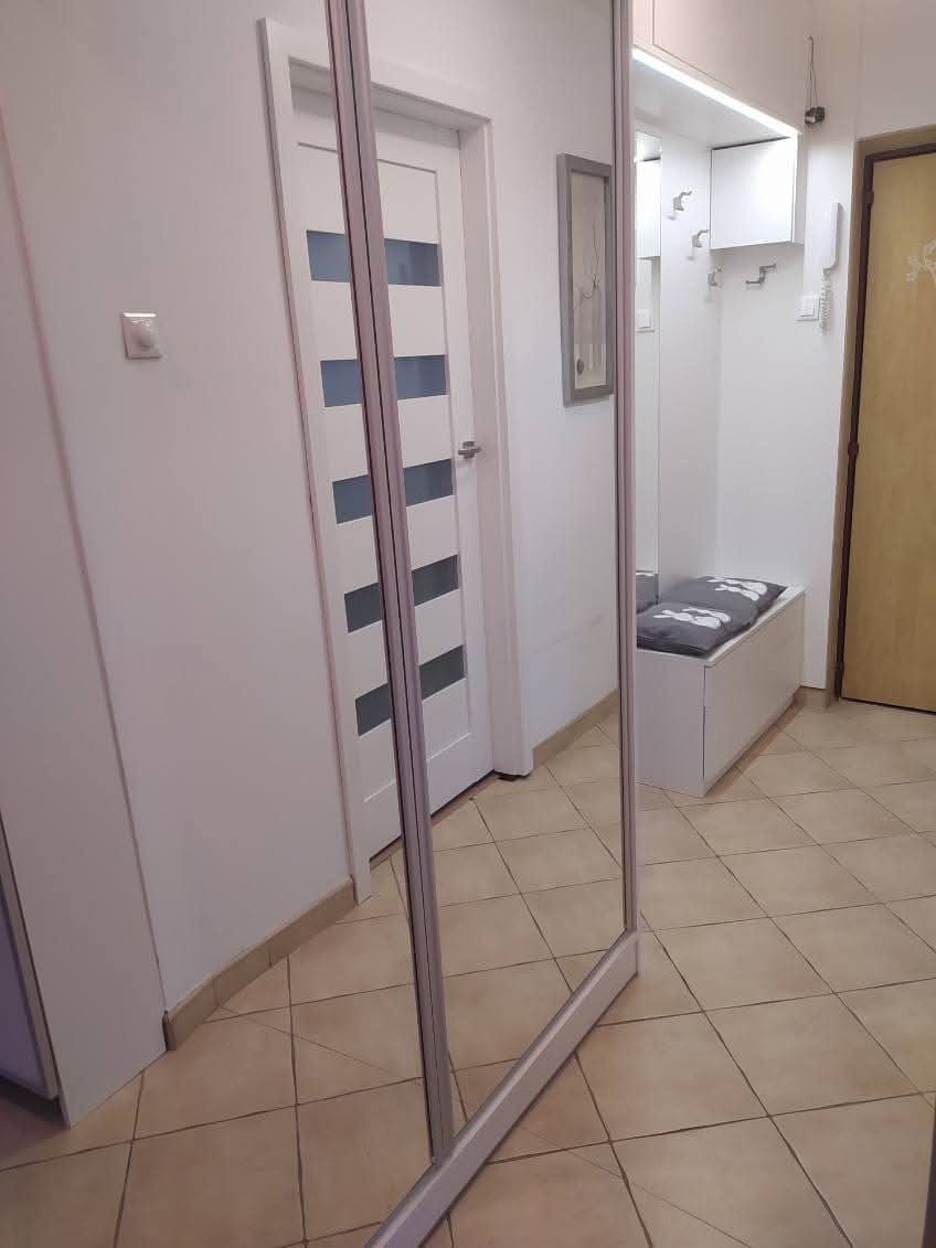 Korytarz z dużą szafą przesuwną z lustrzanymi drzwiami, odbijającą białe drzwi z szybami i białą zabudowę z siedziskiem.