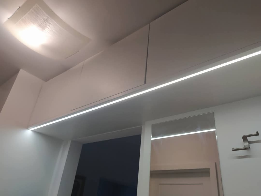 Białe szafki pod sufitem z oświetleniem LED, widoczne odbicie w lustrze i wieszak na ścianie.