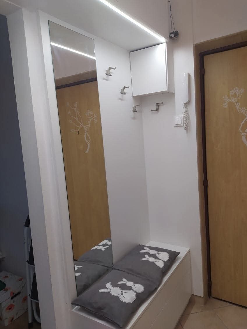 Biała szafa wnękowa z lustrem i siedziskiem z szarymi poduszkami w króliczki, oświetlenie LED podszafkowe, w tle drzwi z dekoracyjnym motywem roślinnym.