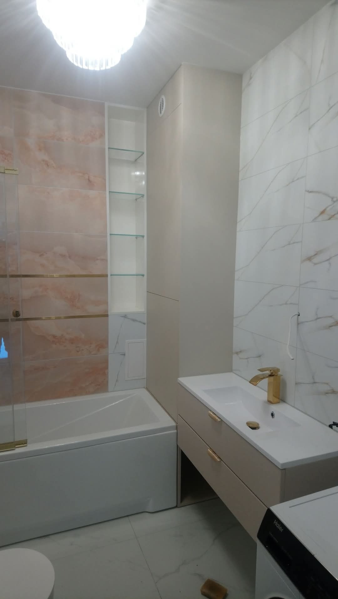 Jasna łazienka z wanną obudowaną płytkami w odcieniach różu, umywalką z szafką z dwoma szufladami z ozdobnymi uchwytami oraz wysoką szafką z półkami z przezroczystego szkła.