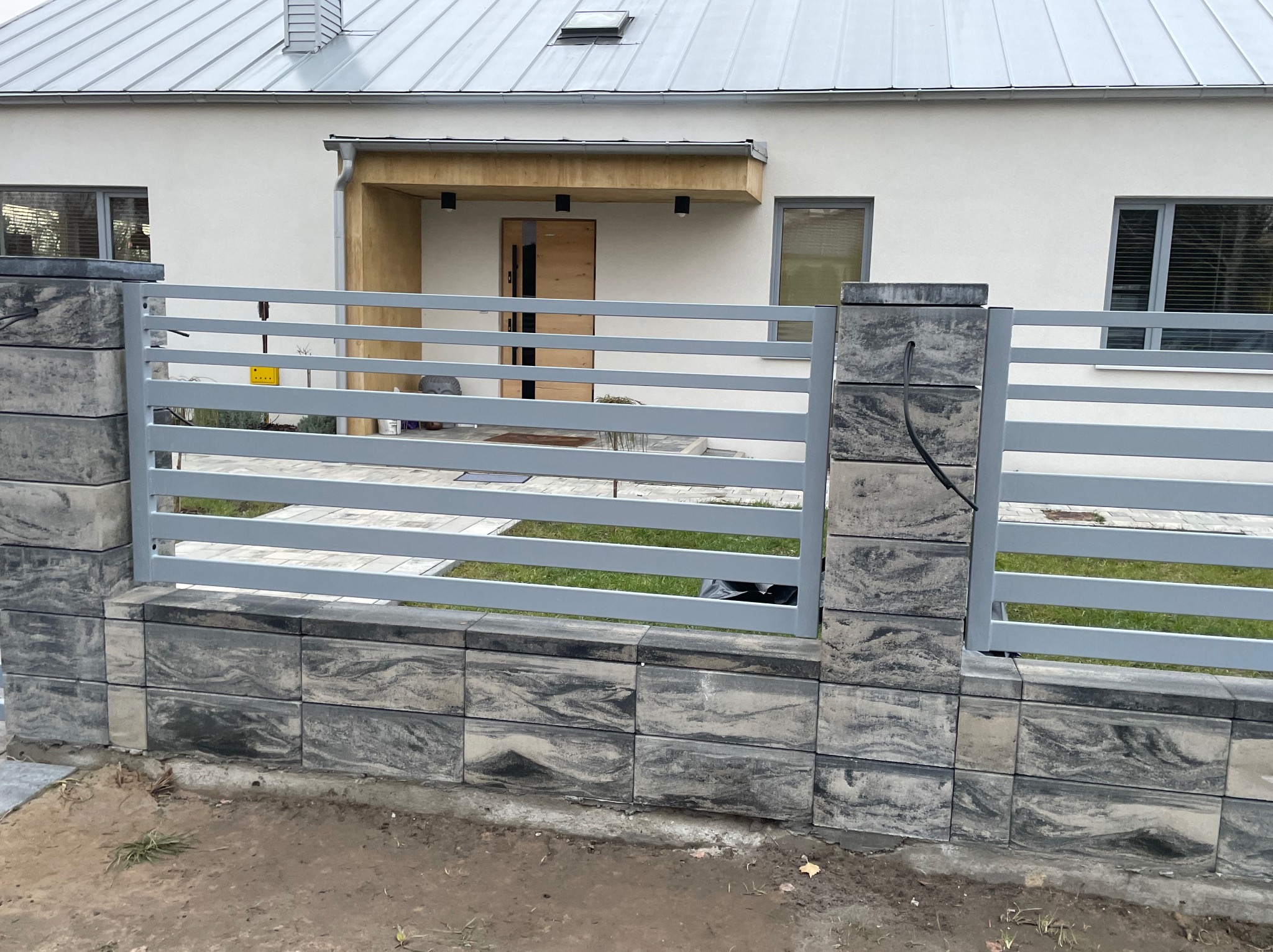 Nowoczesne ogrodzenie panelowe w kolorze szarym z podmurówką z kamienia łupanego na tle domu z szarą blachodachówką.