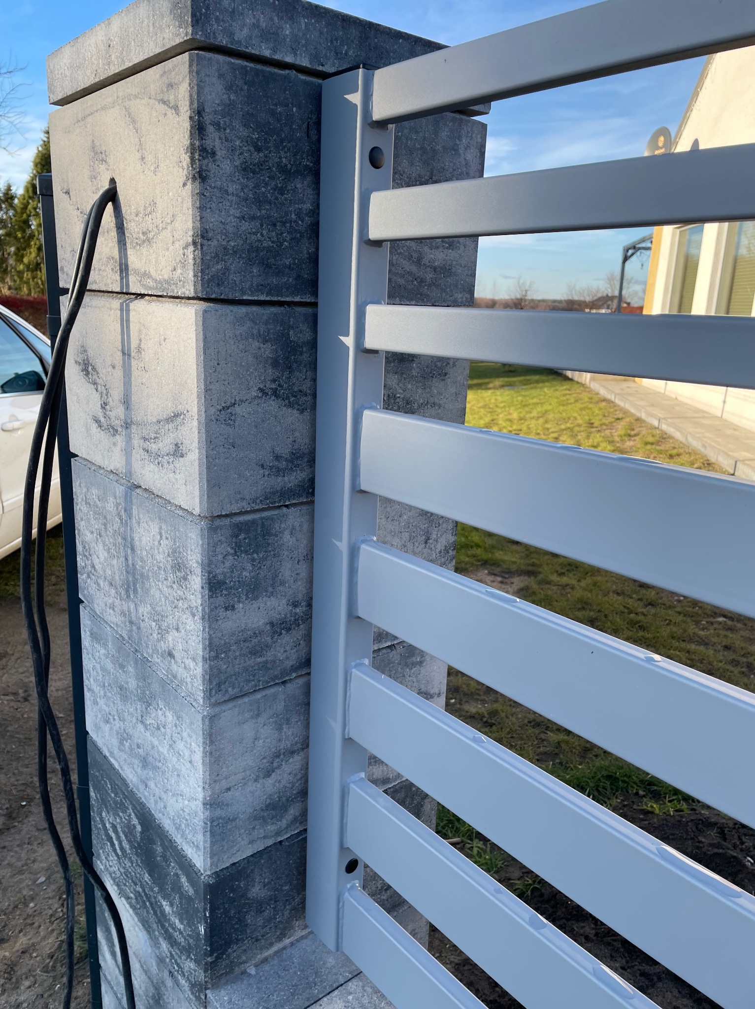 Fragment ogrodzenia panelowego z szarymi, poziomymi przęsłami, zamontowanego na słupku z szarej kostki, z widocznym okablowaniem i fragmentem białego samochodu w tle.