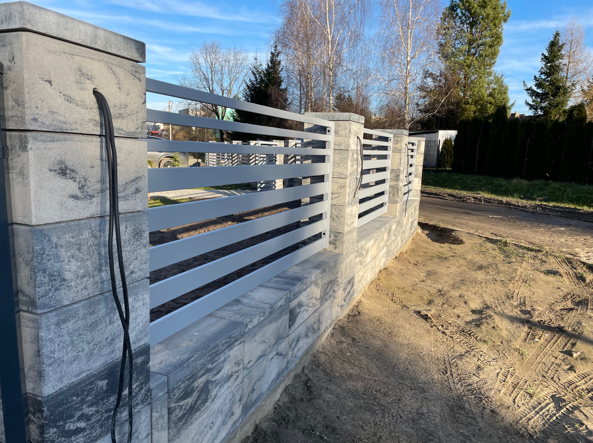 Nowoczesne ogrodzenie panelowe z szarego metalu, osadzone na betonowych słupkach i podmurówce z widocznymi przewodami elektrycznymi wychodzącymi ze słupka. W tle drzewa i niebo.