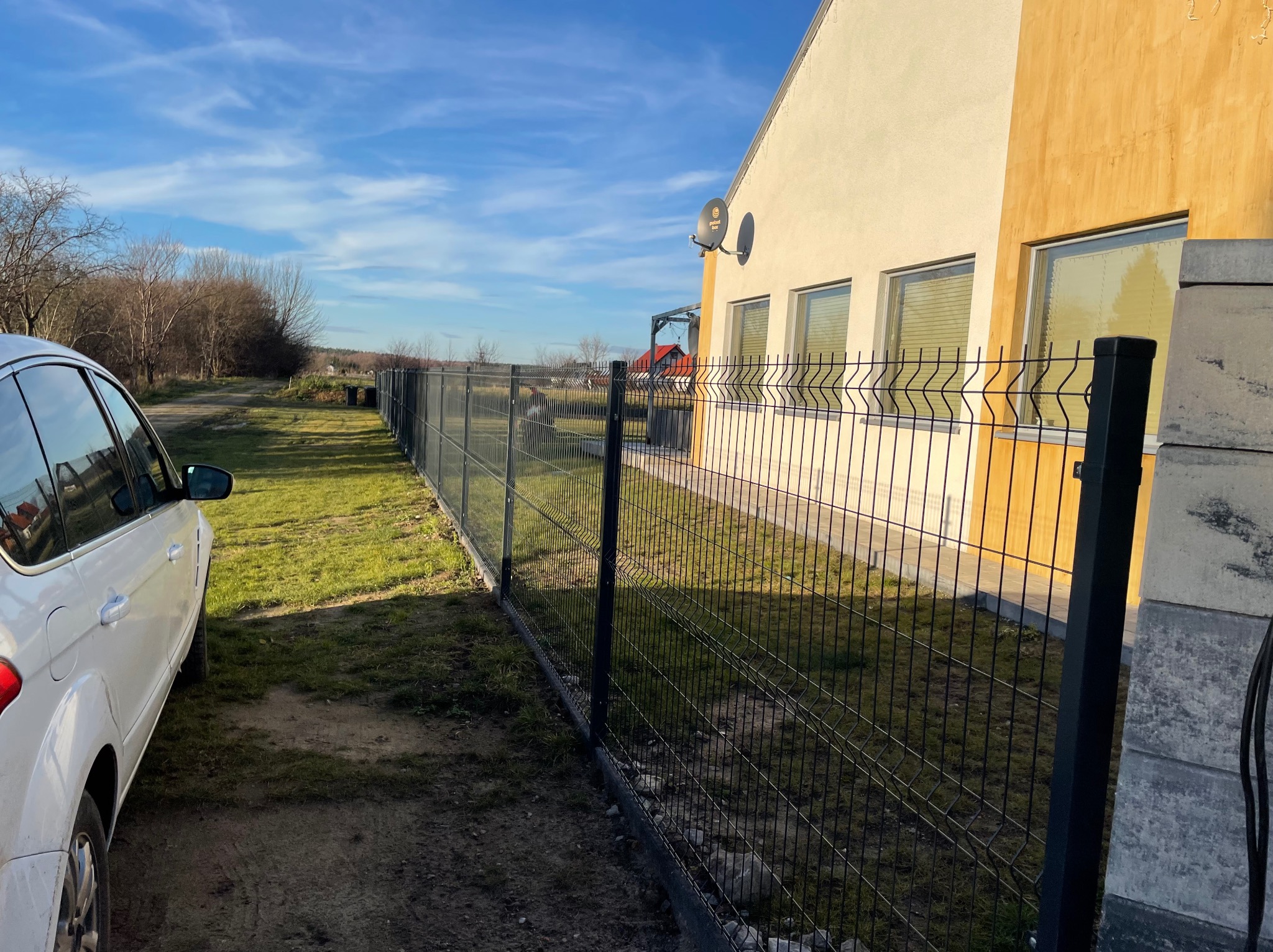 Nowoczesne ogrodzenie panelowe w kolorze grafitowym, z widocznym fragmentem białego samochodu i elewacją domu w odcieniach beżu i żółci, na tle błękitnego nieba z delikatnymi chmurami.