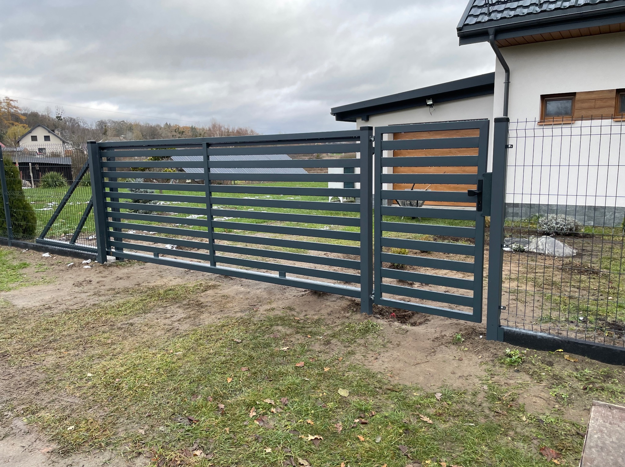 Nowoczesna, pozioma brama wjazdowa w kolorze grafitowym, z poprzecznymi panelami, zamontowana przy domu z jasną elewacją i ciemną dachówką, widoczna trawa z resztkami śniegu.