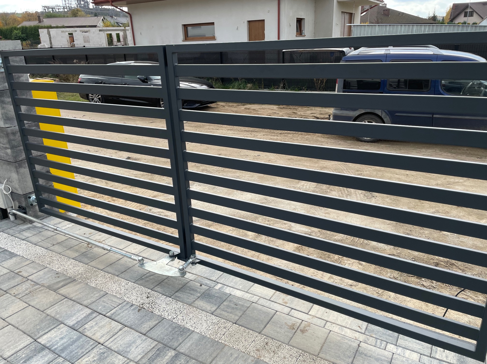 Automatyczna brama wjazdowa z poziomych, stalowych profili, widoczny mechanizm otwierania, szara kostka brukowa, w tle zabudowa jednorodzinna.