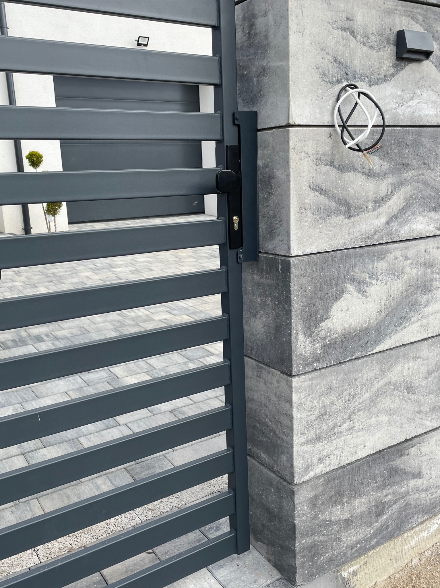 Nowoczesna brama panelowa w kolorze grafitowym z zamkiem, zamontowana na słupkach z szarej kostki betonowej z widocznymi przewodami elektrycznymi do oświetlenia.