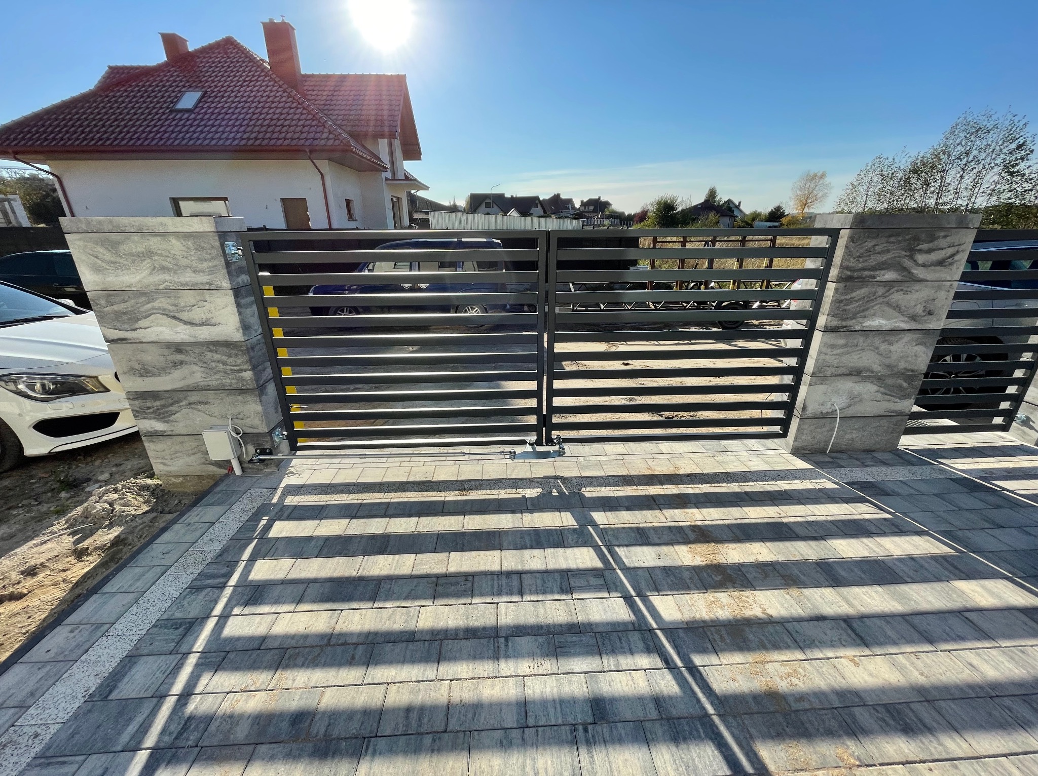 Nowoczesna brama wjazdowa z poziomymi stalowymi profilami, osadzona na słupkach z szarej kostki imitującej kamień, z automatyką, widok z zewnątrz na tle domu z czerwoną dachówką.