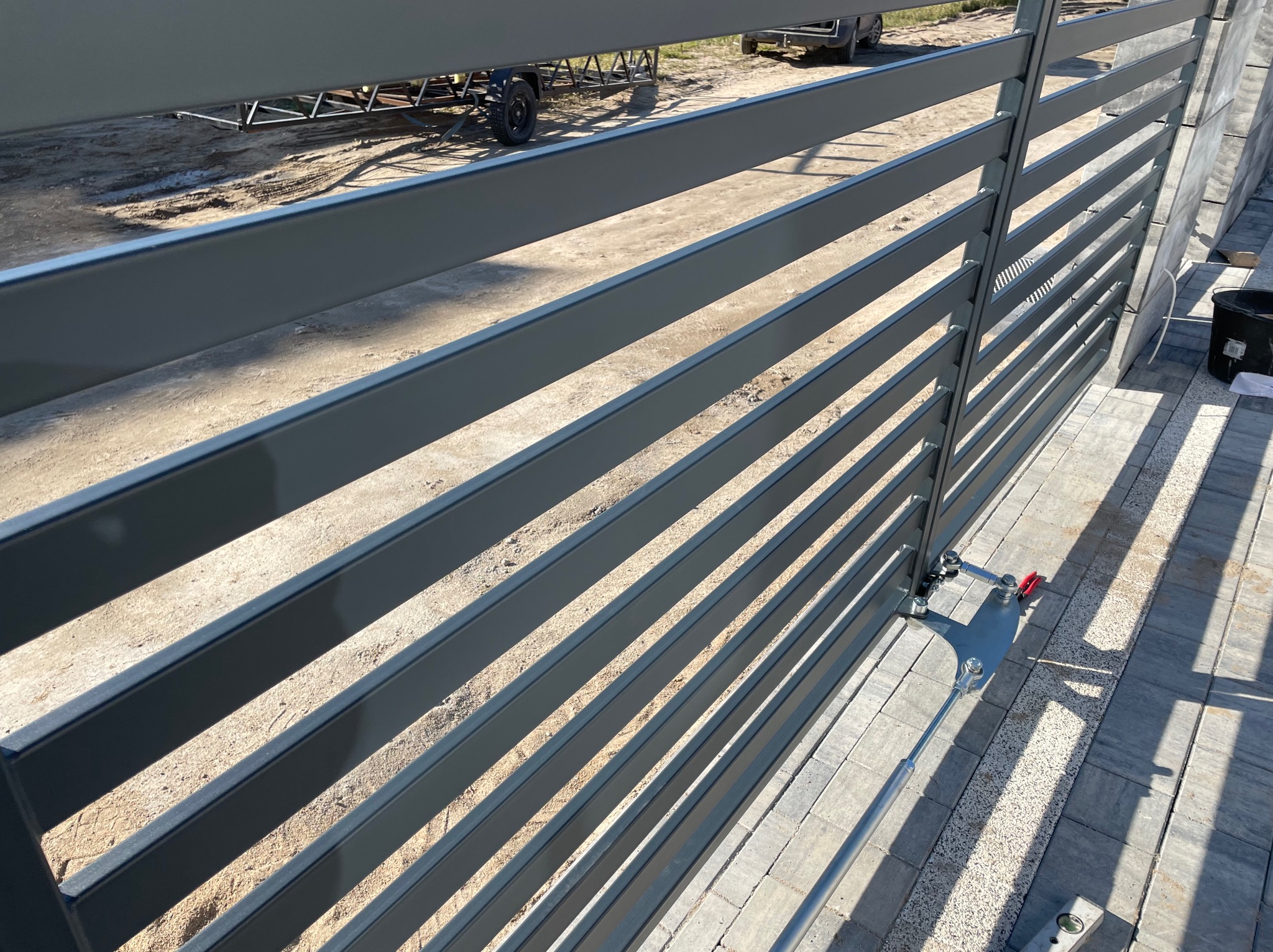 Szara brama przesuwna z poziomymi profilami, widoczny mechanizm automatycznego otwierania, kostka brukowa ułożona na podjeździe, w tle przyczepka samochodowa.