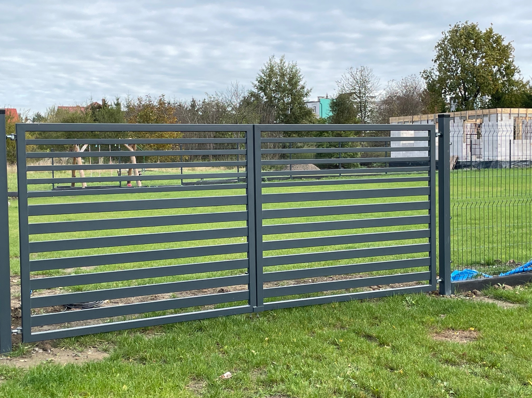 Nowoczesna, pozioma brama wjazdowa z metalowymi panelami w kolorze grafitowym, zamontowana w Mroczno, na tle trawnika i budowy domu.