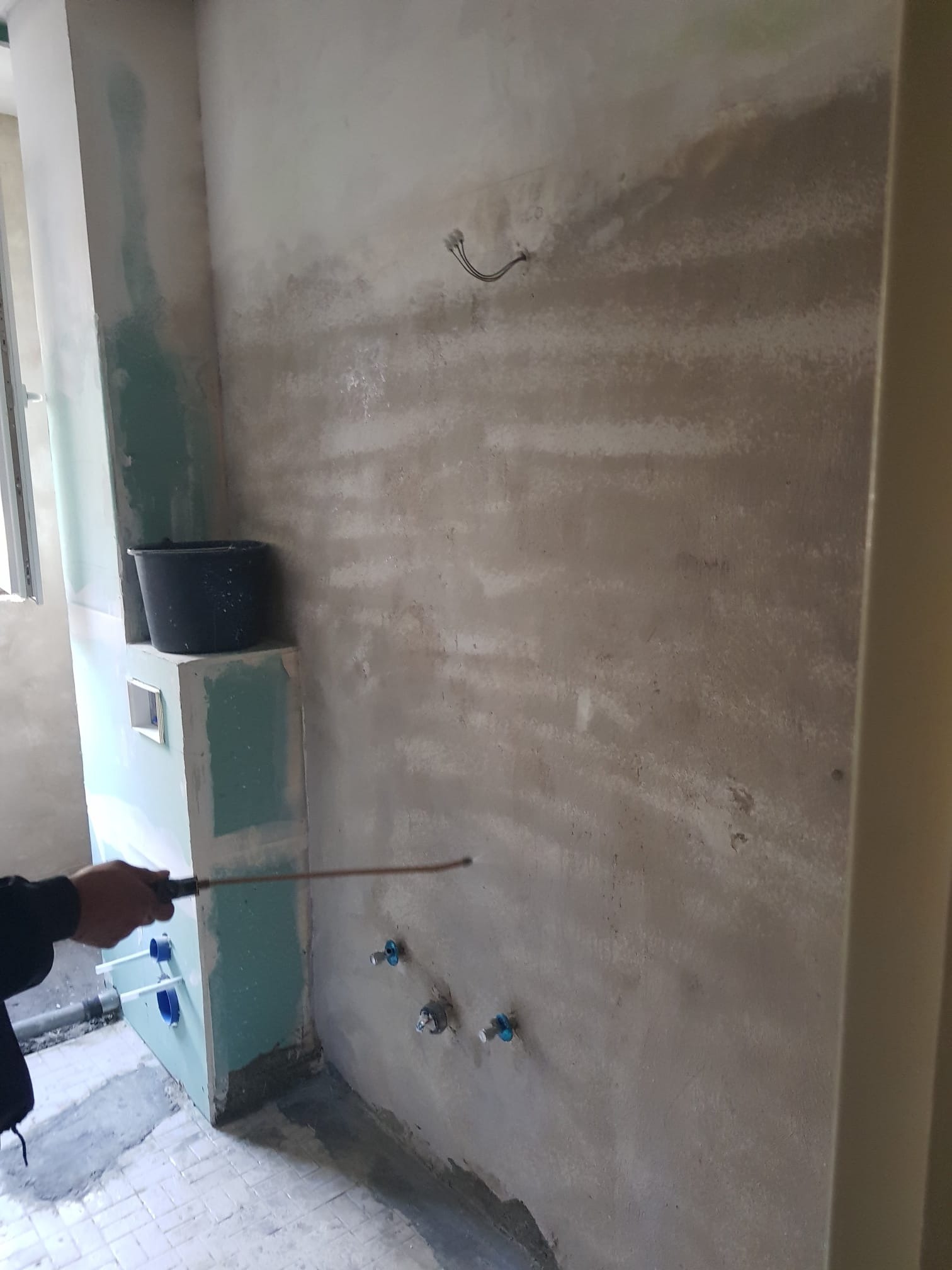 Przygotowanie ściany pod wykończenie w łazience: surowa ściana z widocznymi przyłączami hydraulicznymi, częściowo pokryta gładzią, obok zabudowa z płyty gipsowej z otworem na instalację, czarne...