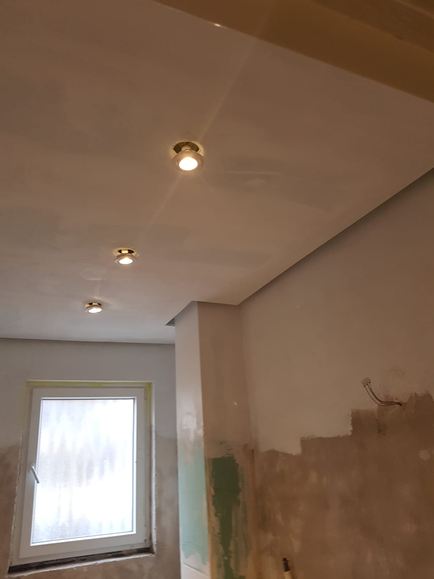 Pokój w trakcie prac wykończeniowych: sufit z zamontowanymi trzema punktowymi lampami, ściany częściowo pomalowane, widoczne przewody elektryczne, okno z białymi ramami.