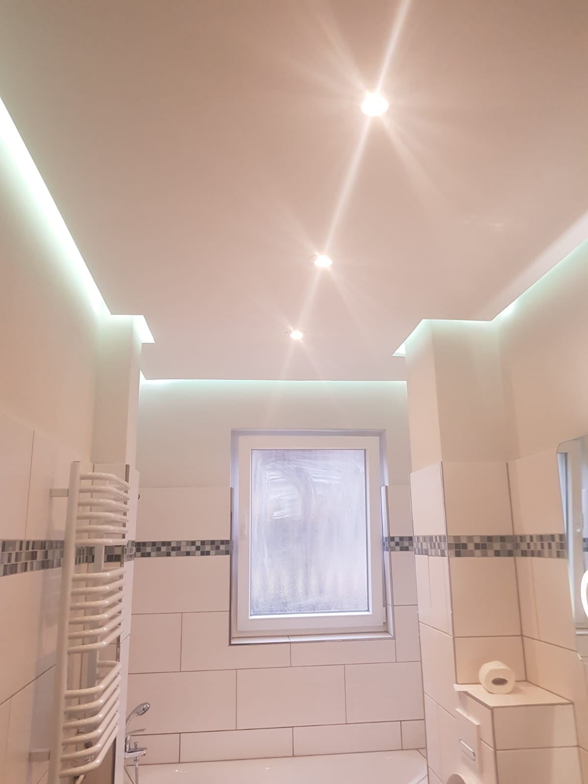 Wykończona łazienka w Sosnowcu z białymi płytkami, podświetlanym sufitem z trzema punktowymi lampami, grzejnikiem drabinkowym i oknem z widocznym osadem.