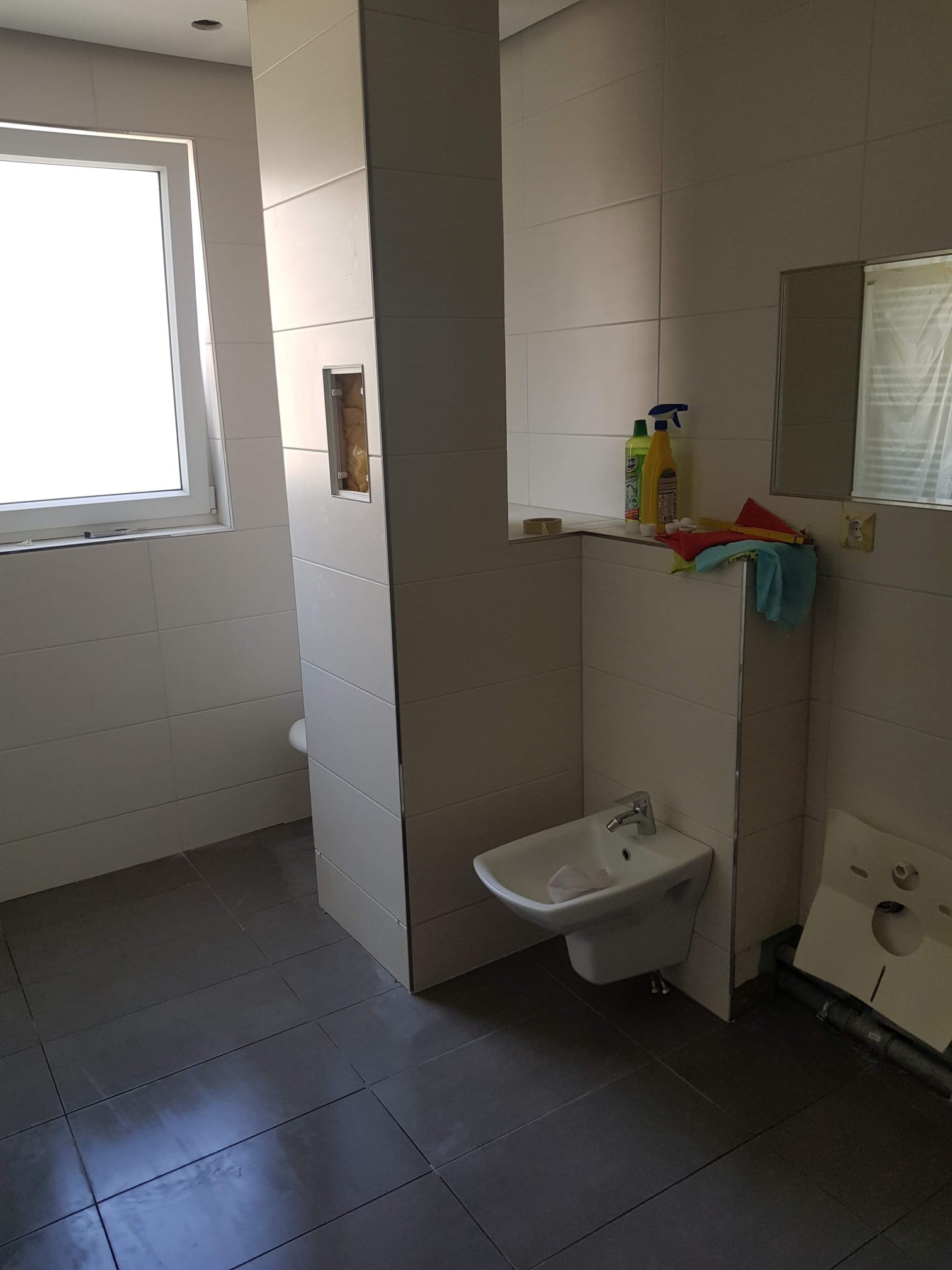 Niedokończona łazienka z szarą podłogą z płytek, białą umywalką bidetową i częściowo obudowaną toaletą podtynkową, widoczne narzędzia i chemia gospodarcza.