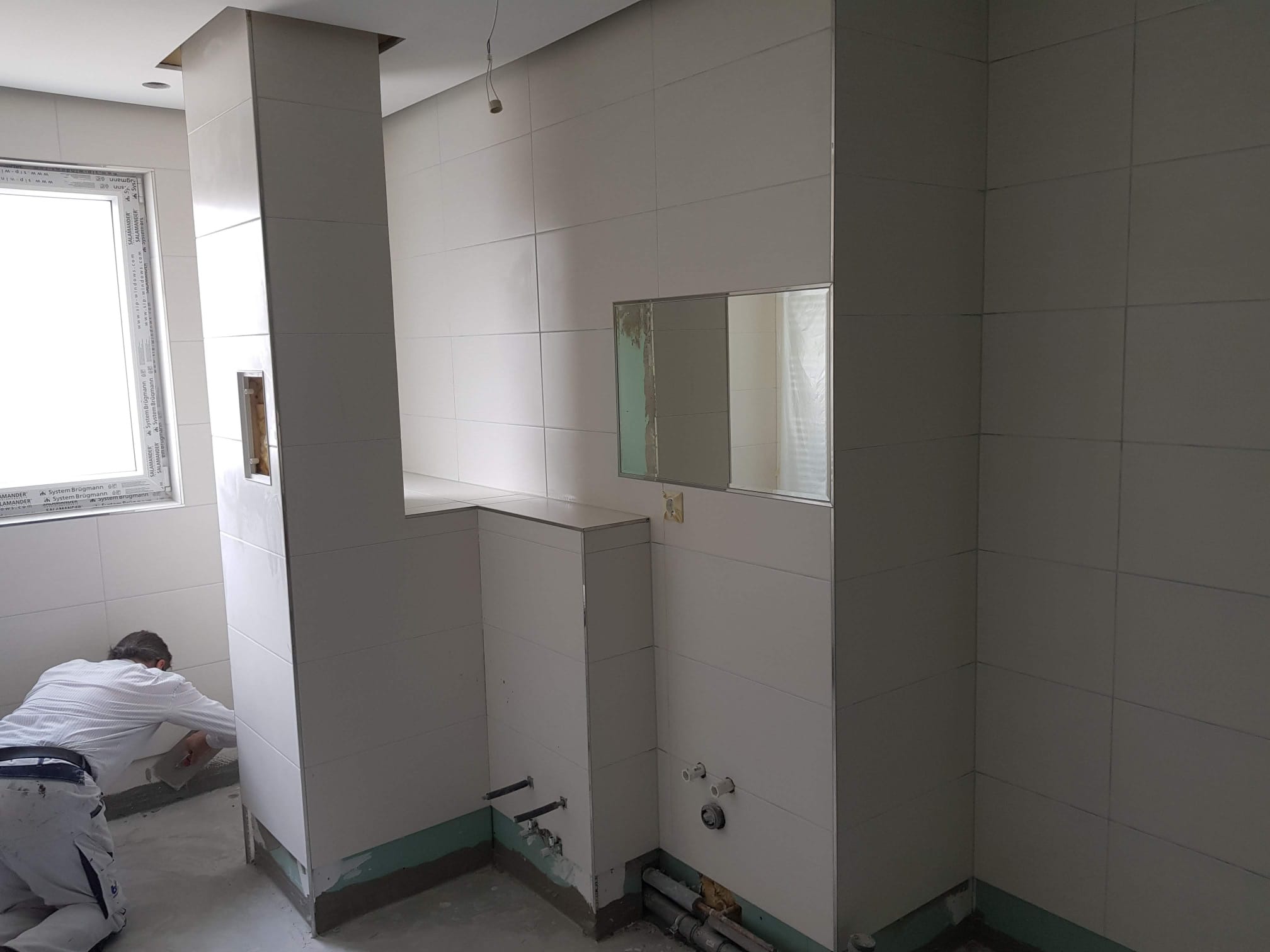 Pracownik w białym kombinezonie wyrównuje ścianę w łazience wyłożonej jasnymi, kwadratowymi płytkami, widoczne niezamaskowane rury i okno z naklejkami producenta.