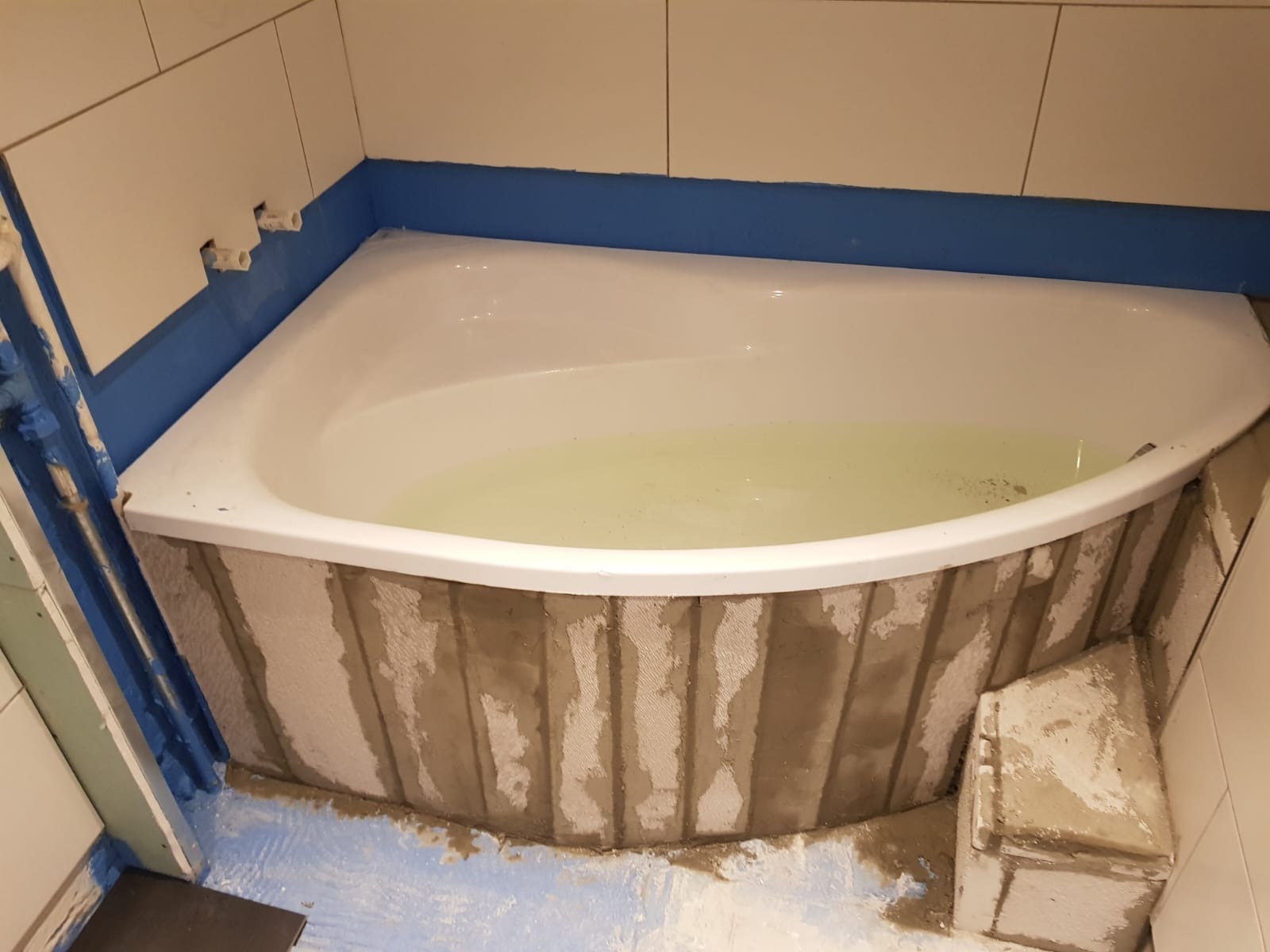 Narożna wanna wypełniona wodą, obudowana surowym betonem z widocznymi śladami po szalunku, zabezpieczona niebieską folią w płynie, w trakcie prac wykończeniowych łazienki.