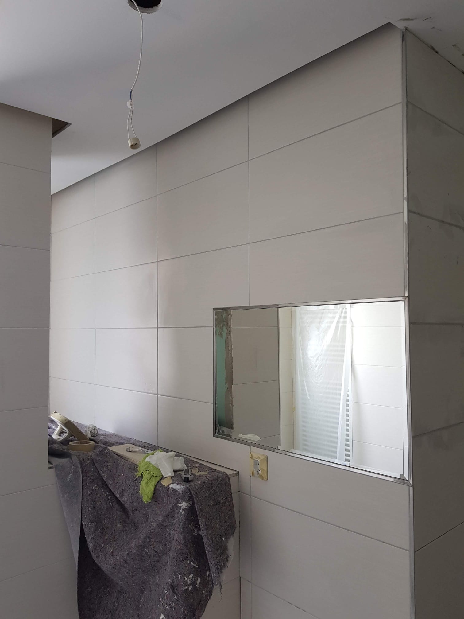 Łazienka w trakcie remontu: białe płytki ścienne z wąskimi fugami, wnęka z lustrem, zabezpieczone narzędzia i materiały na blacie pod ścianą, widoczne niedokończone prace przy suficie.