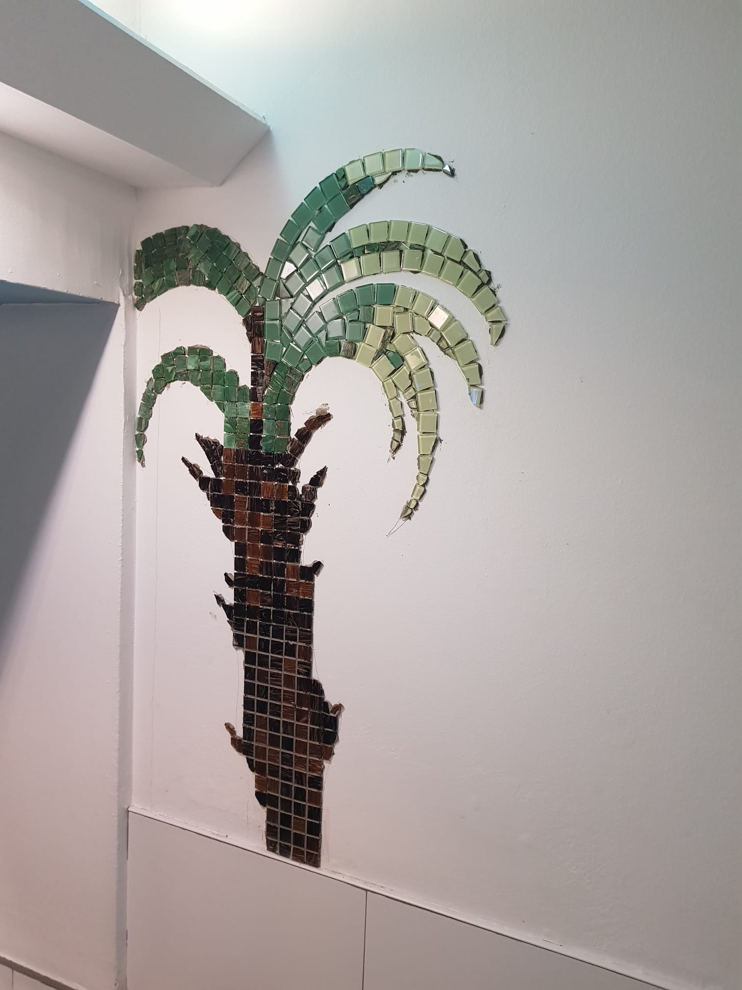 Ściana z dekoracyjną mozaiką w kształcie palmy, wykonana z zielonych i brązowych kwadratowych płytek, częściowo niedokończona, widoczna na białym tle ściany wewnątrz pomieszczenia.